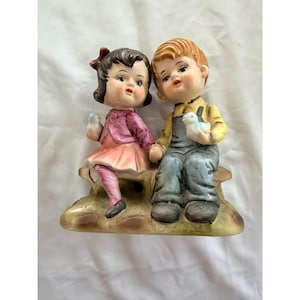 May include: A vintage ceramic figurine depicting a boy and girl seated together on a bench. The girl wears a pink dress and holds a blue bird. The boy wears overalls and a yellow shirt, also holding a blue bird. The figurine has a glossy finish.