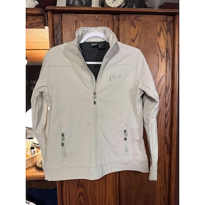 May include: A light beige jacket with a full zipper closure, two side pockets, and a stand-up collar. The jacket has the text "A Paris" embroidered on the front. The jacket is hanging on a white hanger.