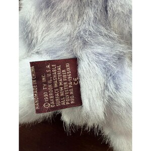 May include: Close-up of a plush toy with a tag. The toy has a soft, fluffy texture in shades of light blue and white. The tag is a dark red color and includes text such as "1993 TY INC." and "SURFACE WASHABLE."