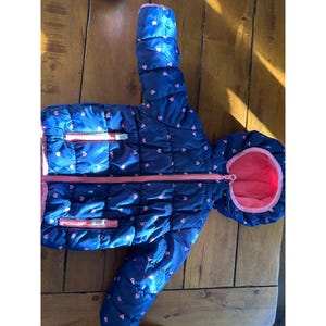 May include: A navy blue quilted jacket with a hood, featuring a pink heart pattern. The jacket has a pink interior and pink zips. The jacket is designed for cold weather and is likely for a child.