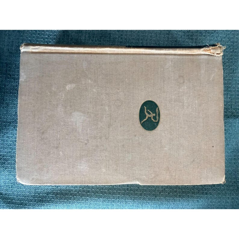 May include: A vintage book with a light tan cover and a green oval emblem with Arabic script. The spine is a lighter color, and the edges show wear. The book is rectangular and appears to be closed.