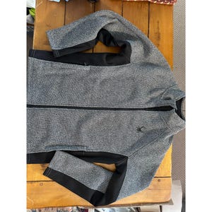 May include: A gray and black zip-up jacket with a small logo on the chest. The jacket has black panels on the sides and sleeves. The jacket is made of a textured material and has a full-length zipper.