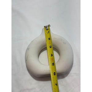 May include: A white, donut-shaped ceramic vase with a narrow neck. A yellow measuring tape shows the vase is approximately 6 inches in diameter. The vase has a smooth, matte finish and a circular opening in the center.