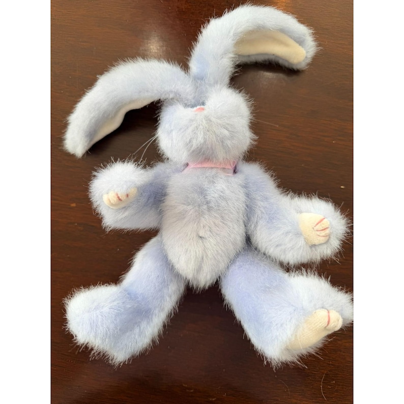 May include: A plush, light blue bunny with long ears and a pink ribbon around its neck. The bunny has a soft, fluffy texture, with cream-colored accents on its paws and the inside of its ears. A charming stuffed animal.