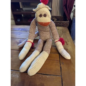 May include: A handmade sock monkey doll with a beige hat and a red pom-pom. The monkey has brown button eyes, an orange mouth, and red accents on its arms. The body and arms are brown and white, while the legs are white.