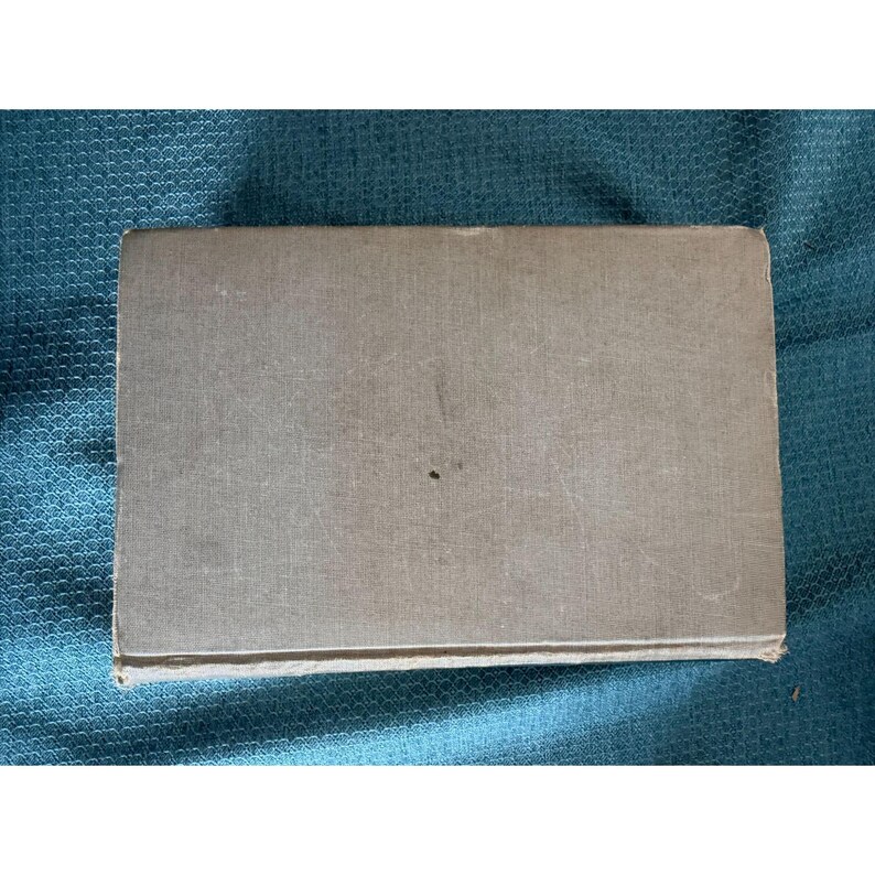 May include: A rectangular, light gray book with a textured cover. The book has a small, dark spot on the front cover. The edges of the book are slightly worn, suggesting it is vintage or has been well-used. The book is lying on a teal-colored surface.
