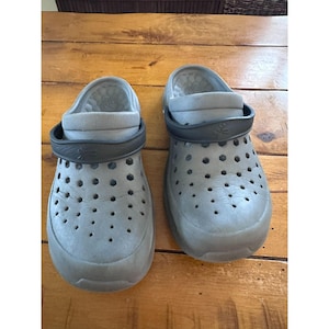May include: A pair of gray clogs with numerous small holes for ventilation. The clogs have a rounded toe and a black strap that can be worn around the heel. The shoes are made of a lightweight material.