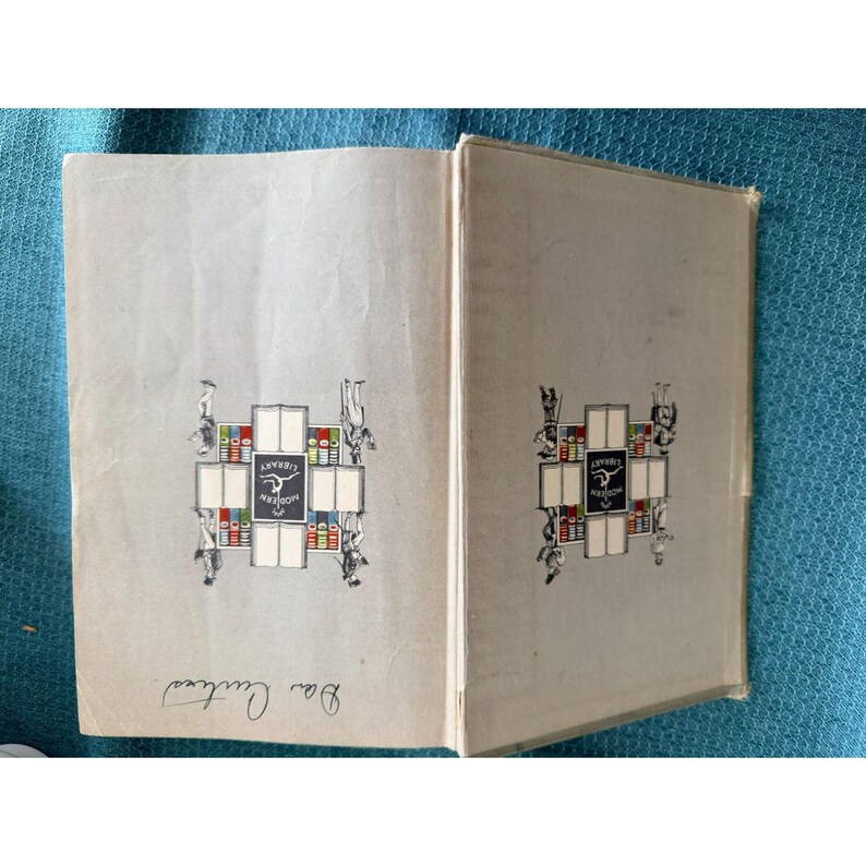 May include: An open book with light gray pages. The cover and inside pages feature a decorative emblem with the words "Modern Library" and illustrations of figures and books. The book is open to reveal the title page.
