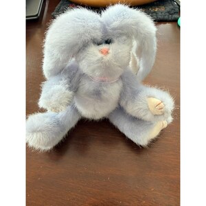 May include: A plush, lavender-colored bunny rabbit with long, floppy ears and a pink nose. The stuffed animal has a light pink ribbon around its neck and white accents on its paws and ears. The bunny is sitting on a wooden surface.