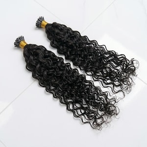 Puede incluir: Dos paquetes de extensiones de cabello rizado oscuro. El cabello es de color negro intenso con un patrón de ondas natural. Los extremos de los paquetes están asegurados con una banda dorada. Las extensiones están dispuestas sobre una superficie blanca.