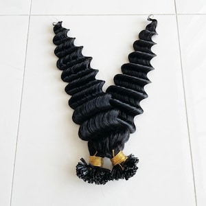 Puede incluir: Dos mechones de extensiones de cabello negro con una textura ondulada. Las extensiones están atadas en la base con alambre dorado. El cabello está dispuesto en forma de V sobre un fondo blanco.