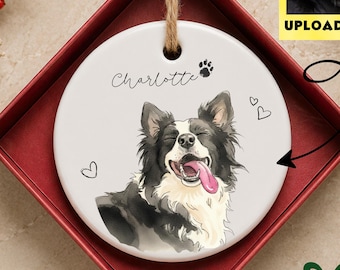 Puede incluir: Un adorno redondo de cerámica blanca con una ilustración de un perro blanco y negro con la lengua rosa, el nombre Charlotte, una huella y corazones pequeños. Cuelga de un cordel de yute dentro de una caja de regalo roja cuadrada.