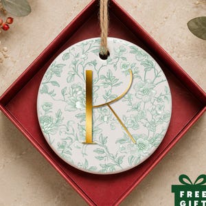May include: A round white ceramic ornament featuring a light green floral pattern and a large gold letter K in the center. The ornament hangs from a brown twine loop and is presented inside a square red gift box with a green gift box icon.