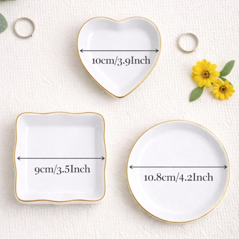 May include: Three white ceramic trinket dishes with gold rims. The dishes are heart-shaped (3.9 inches), square (3.5 inches), and round (4.2 inches). The dimensions are indicated on the dishes. The background is a white surface with small flowers and rings.
