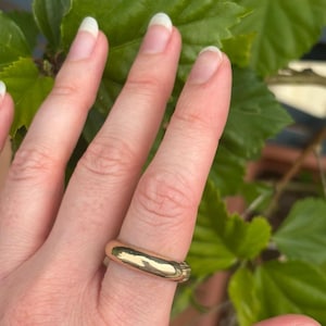 Puede incluir: Un anillo de banda en tono dorado pulido con una superficie lisa y redondeada. El anillo presenta una construcción simple y sólida con un acabado metálico reflectante, usado en un dedo sobre un fondo de hojas verdes.