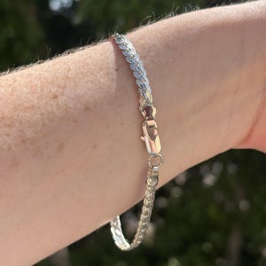 May include: A silver-toned chain bracelet featuring a flat, braided link design with a polished finish. The bracelet is secured with a lobster claw clasp, which connects two ends of the woven metal chain, creating a continuous, flexible loop.