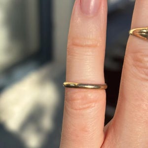 May include: A simple, polished gold band ring with a smooth, rounded profile. The ring is worn on a finger, showcasing its minimalist design and reflective metallic surface. A second, partially visible gold ring is positioned on an adjacent finger.