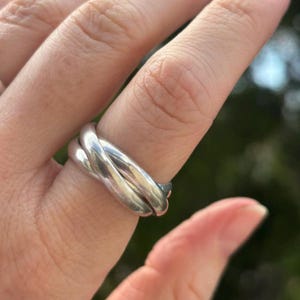 May include: A polished silver-toned ring featuring three interlocking bands that overlap to create a continuous, rounded shape. The metal surface is smooth and reflective, showing subtle highlights and shadows across the curved, interconnected loops.
