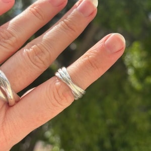 May include: A polished silver ring featuring a triple band design that crosses over itself in the centre. The metal has a smooth, reflective surface with a continuous, flowing appearance that wraps around the finger in a layered, intertwined style.