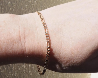 Puede incluir: Una pulsera de cadena figaro en tono dorado fino que presenta un patrón repetitivo de tres eslabones circulares pequeños seguidos de un eslabón ovalado alargado. La cadena metálica descansa sobre la piel, mostrando un acabado pulido y un diseño clásico.