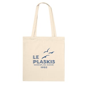 Puede incluir: Bolso tote de color crema con asas largas. El bolso presenta el texto "LE PLASKIS MARIAPLAN, SUEDE 1932" en azul marino, junto con un gráfico de tres pájaros volando. El bolso está hecho de una tela resistente.