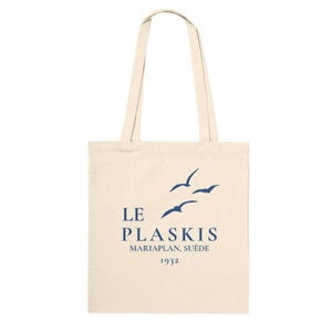 May include: A cream-colored canvas tote bag with two long shoulder straps. The front features a dark blue graphic of three flying birds above the text LE PLASKIS, MARIAPLAN, SUÈDE, and the year 1932 printed in a clean, dark blue serif font.