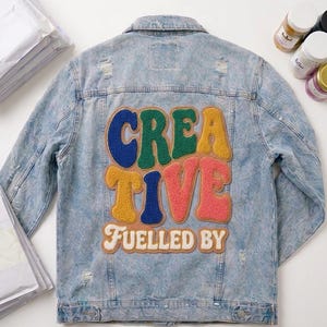 Puede incluir: Chaqueta vaquera azul claro con diseño desgastado. La parte trasera presenta la palabra "CREATIVE" en letras coloridas y texturizadas, con "FUELLED BY" debajo. Una prenda de vestir elegante y artística.