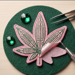 May include: A pink leaf-shaped patch embellished with green rhinestones, placed on a dark green felt circle. Two loose green faceted gems, a pair of metal tweezers, and a metal stylus tool are positioned around the patch.