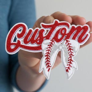 Op de afbeelding: Een rood en wit honkbal-thema patch met het woord "Custom" in een scriptlettertype. De patch heeft een honkbal strik ontwerp met rode stiksels en zilveren glitter accenten. De patch wordt in de hand van een persoon gehouden.