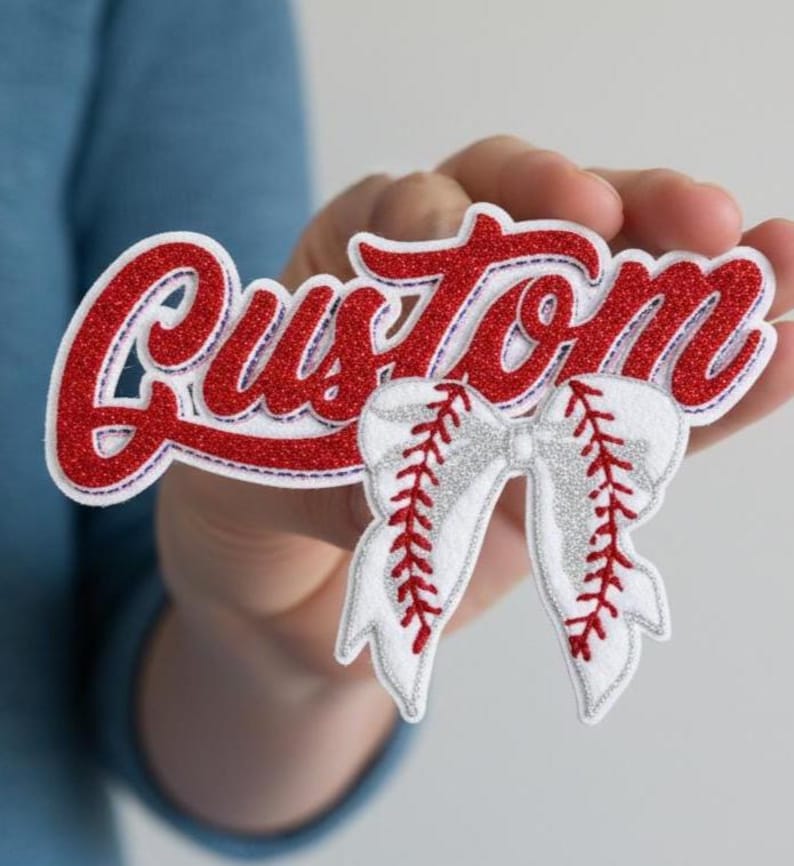 Puede incluir: Un parche rojo y blanco con tem&aacute;tica de b&eacute;isbol con la palabra "Custom" en una fuente de escritura. El parche presenta un dise&ntilde;o de lazo de b&eacute;isbol con costuras rojas y detalles de purpurina plateada. El parche se sostiene en la mano de una persona.