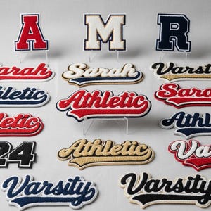 Puede incluir: Surtido de parches universitarios bordados en varios colores y dise&ntilde;os. Incluye letras, nombres y la palabra "Athletic" en rojo, azul y dorado. Los parches se muestran sobre soportes transparentes sobre un fondo claro.