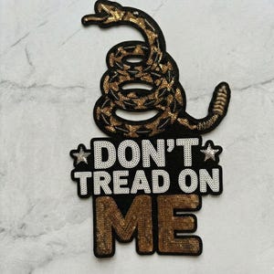 May include: A sequined patch featuring a coiled rattlesnake above the phrase "DON'T TREAD ON ME." The snake is brown and gold, and the text is white and gold on a black background. The patch is likely for clothing or accessories.