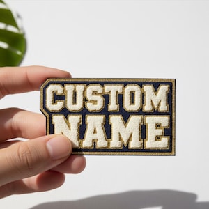 Puede incluir: Un parche azul marino y crema con las palabras "CUSTOM NAME" en una fuente audaz de estilo universitario. El parche tiene un borde dorado y es de forma rectangular.