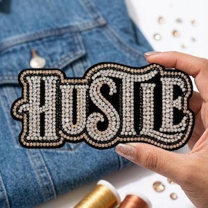Puede incluir: Un parche negro con la palabra "HUSTLE" escrita con brillantes pedrería. El parche tiene un borde dorado y se muestra sobre una chaqueta vaquera. El parche es un artículo decorativo.