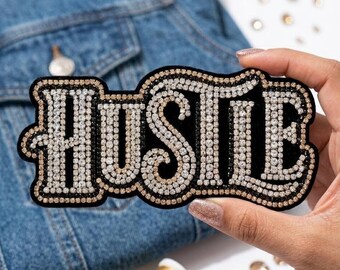 May include: A black patch with the word "HUSTLE" spelled out in sparkling rhinestones. The patch has a gold border and is held up against a denim jacket. The patch is a decorative item.