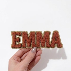 Peut inclure: Un écusson marron texturé avec le nom "EMMA" en majuscules. Les lettres ont une texture floue et sont bordées d'or. L'écusson est tenu sur un fond blanc.
