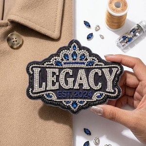 Puede incluir: Un parche azul marino con la palabra "LEGACY" en pedrería plateada, enmarcado por un borde negro con adornos azules y plateados. El texto "EST. 2024" está debajo. También se ve una bobina de hilo dorado y gemas decorativas.