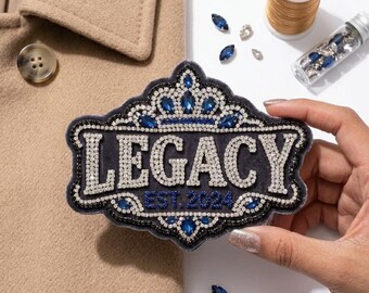 May include: A navy blue patch with the word "LEGACY" in silver rhinestones, framed by a black border with blue and silver embellishments. The text "EST. 2024" is below. A spool of gold thread and decorative gems are also visible.