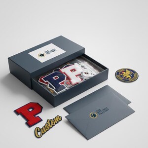 Puede incluir: Una caja gris oscuro con el texto "The Patches Factory" en la tapa, que contiene parches bordados. Los parches incluyen la letra "P" en rojo, blanco y azul, y un parche personalizado con la palabra "Custom".