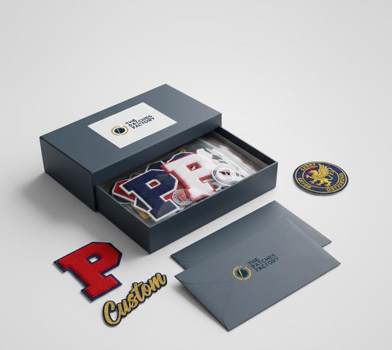 Puede incluir: Una caja gris oscuro con el texto "The Patches Factory" en la tapa, que contiene parches bordados. Los parches incluyen la letra "P" en rojo, blanco y azul, y un parche personalizado con la palabra "Custom".