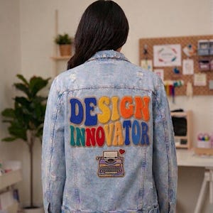 Puede incluir: Chaqueta vaquera azul claro con detalles desgastados. La parte trasera presenta un parche colorido con las palabras "DESIGN INNOVATOR" sobre un diseño de máquina de escribir bordada. Un artículo de moda creativo.