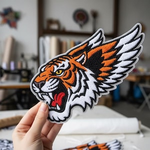 May include: Embroidered tiger patch with orange, black, and white details. The tiger's head is open-mouthed, with a red tongue and sharp teeth. The patch has a wing-like design and a grey border. Suitable for jackets, bags, or other apparel.