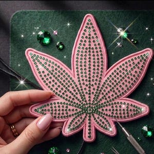 May include: A pink fabric patch shaped like a seven-pointed leaf, densely embellished with small, sparkling green rhinestones arranged in rows. The patch features a stitched pink border and is surrounded by loose green rhinestones.