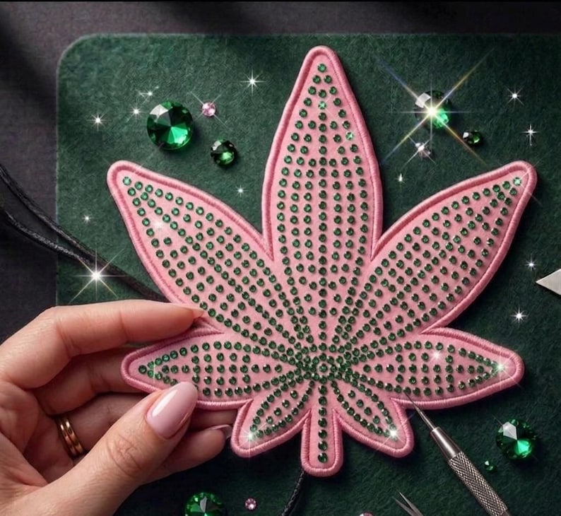May include: A pink fabric patch shaped like a seven-pointed leaf, densely embellished with small, sparkling green rhinestones arranged in rows. The patch features a stitched pink border and is surrounded by loose green rhinestones.