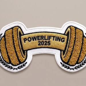 Puede incluir: Parche bordado con un diseño de barra dorada y el texto "POWERLIFTING 2025" en una pancarta dorada. Los extremos de la barra tienen una apariencia texturizada, y el parche está delineado con un borde blanco y hojas de laurel negras.