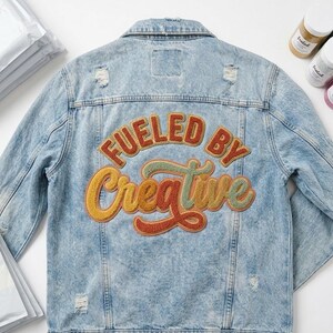 Puede incluir: Chaqueta vaquera azul claro con detalles desgastados. La parte trasera presenta un parche bordado colorido con las palabras "FUELED BY Creative" en naranja, amarillo y verde. La chaqueta tiene cuello clásico, cierre de botones y bolsillos.