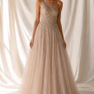 Puede incluir: Un vestido largo de un solo hombro en tono beige champán, con un corpiño ajustado cubierto de lentejuelas densas y brillantes, y una falda de tul amplia y fluida con un brillo sutil en toda la tela, creando una silueta voluminosa y superpuesta.