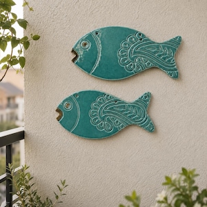 May include: Two teal ceramic fish wall decorations featuring embossed paisley and floral patterns on their bodies. Each fish has a small circular eye detail and an open mouth, with a smooth, glossy finish across the entire surface area.