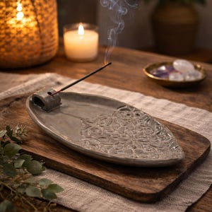 May include: A gray ceramic incense holder with a floral design, holding a lit incense stick with smoke rising. The holder sits on a wooden tray with a candle and other decorative items in the background. A relaxing, spa-like theme.