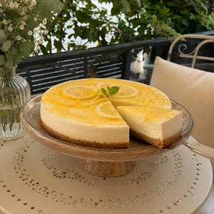 May include: A round lemon cheesecake on a textured pedestal plate, featuring a smooth cream-colored filling, a golden-brown crust, and a bright yellow lemon glaze topping. It is garnished with thin lemon slices and a small sprig of green mint.